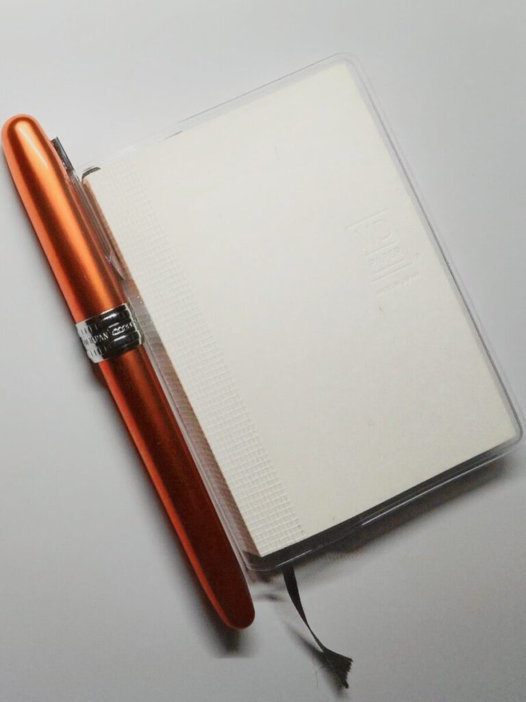 メモ帳のカバーに挿されたプラチナ万年筆プレジール（ノバオレンジ）。実用性と色の存在感が際立つ拡大写真