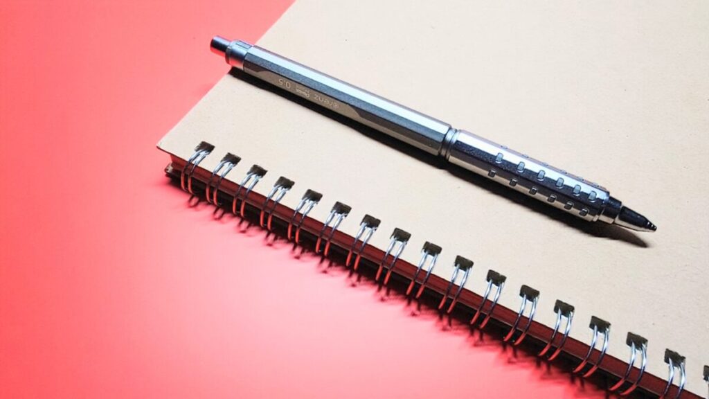 朱色背景のノート表紙に、オレンズATがクリップで留められた構図。軽量で親しみやすいデザインが、学生の日常使いを想起させる写真。